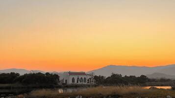 A white building with arched windows, situated by a body of water with mountains in the background.