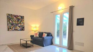 Una sala de estar moderna con sofá, una mesa de centro de vidrio, un cuadro abstracto de colores vivos, una lámpara de pie y un ventanal corredero que da a un espacio exterior.