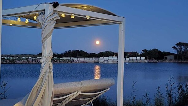 Un espacio al aire libre con asientos blancos, un toldo, luces colgantes y vista de la luna sobre un cuerpo de agua.