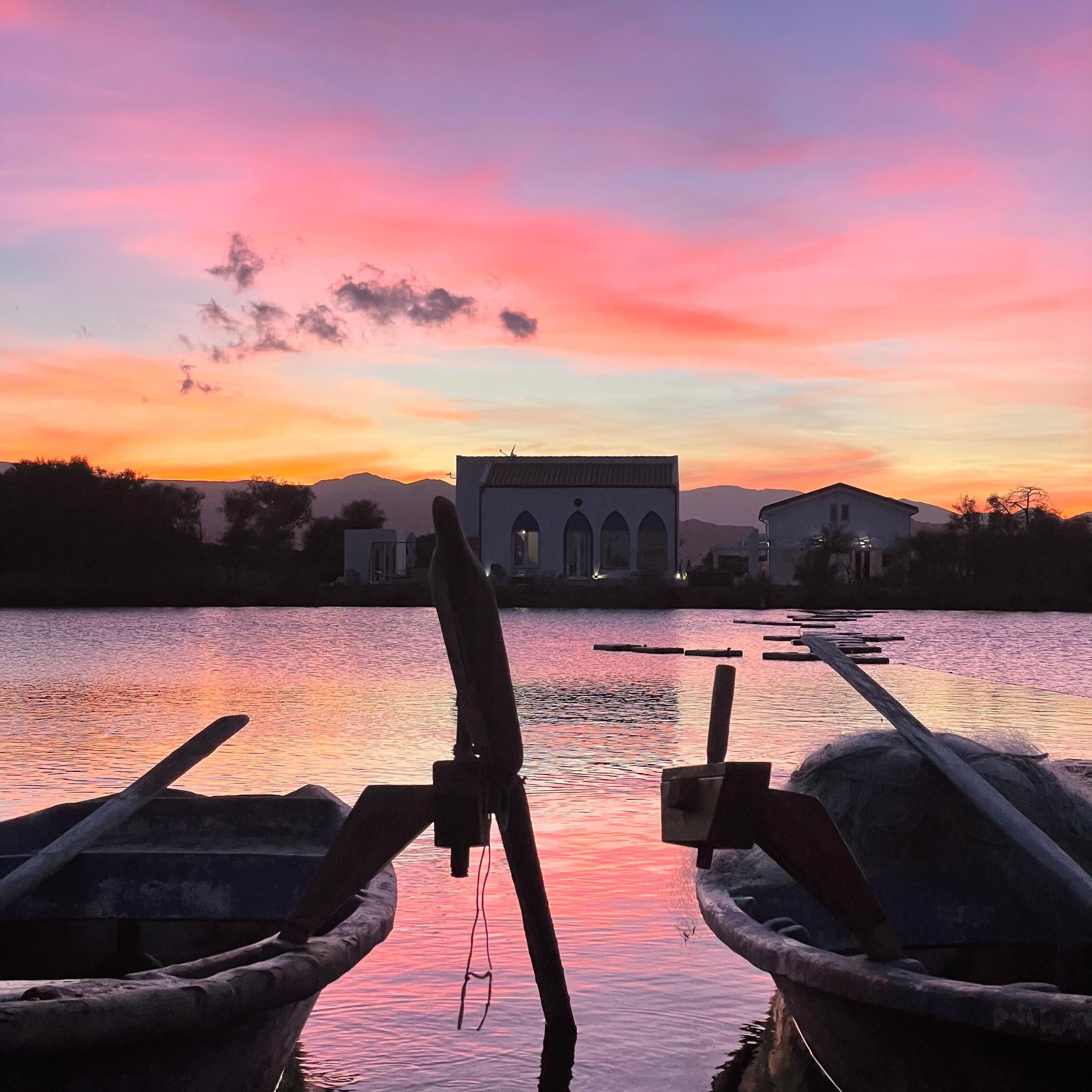 Ein Sonnenuntergang vom Boot aus, mit zwei Booten im Vordergrund und einem Gebäude mit Bögen im Hintergrund.