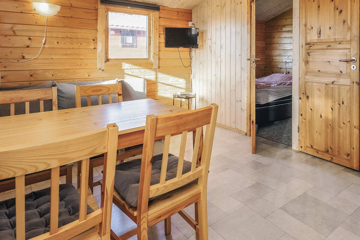 A wooden cabin interior with a dining table, chairs, a television, and a bed.