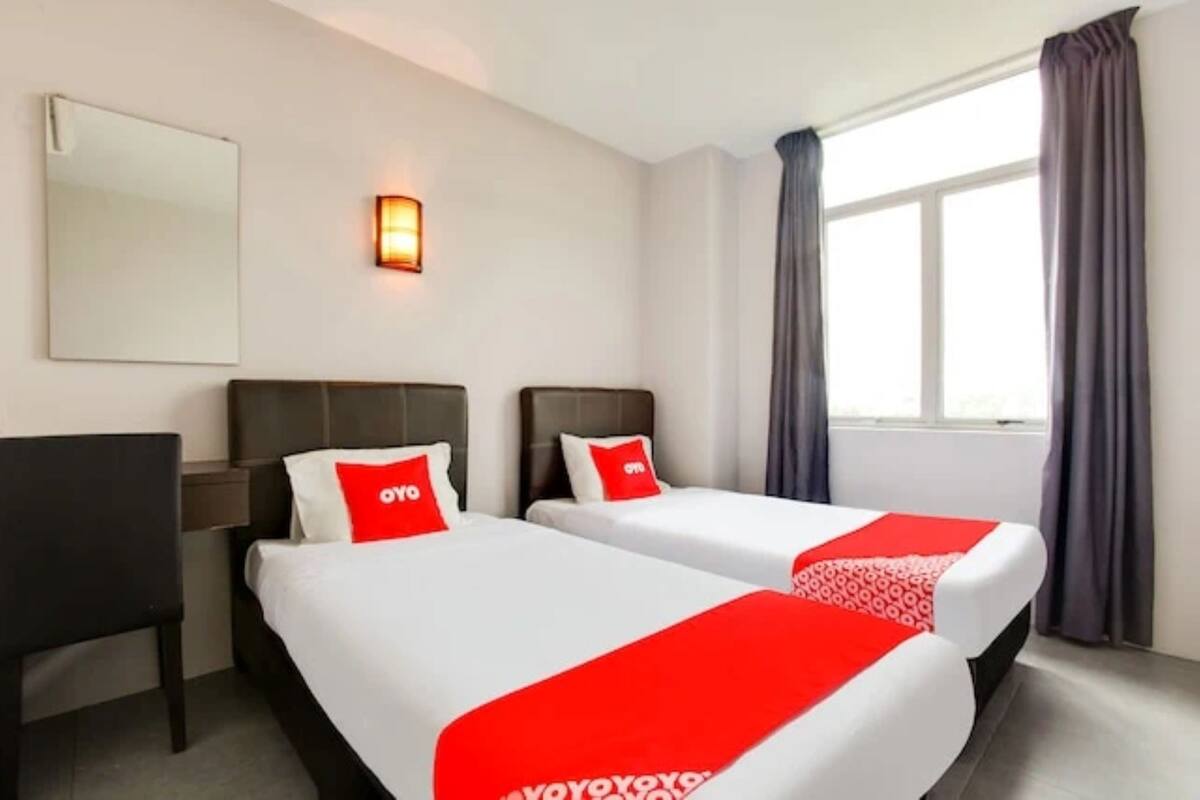 Una camera d'albergo con due letti, ognuno con copripiumino rosso e bianco, un comodino, una sedia, una lampada a parete e una finestra con tende.