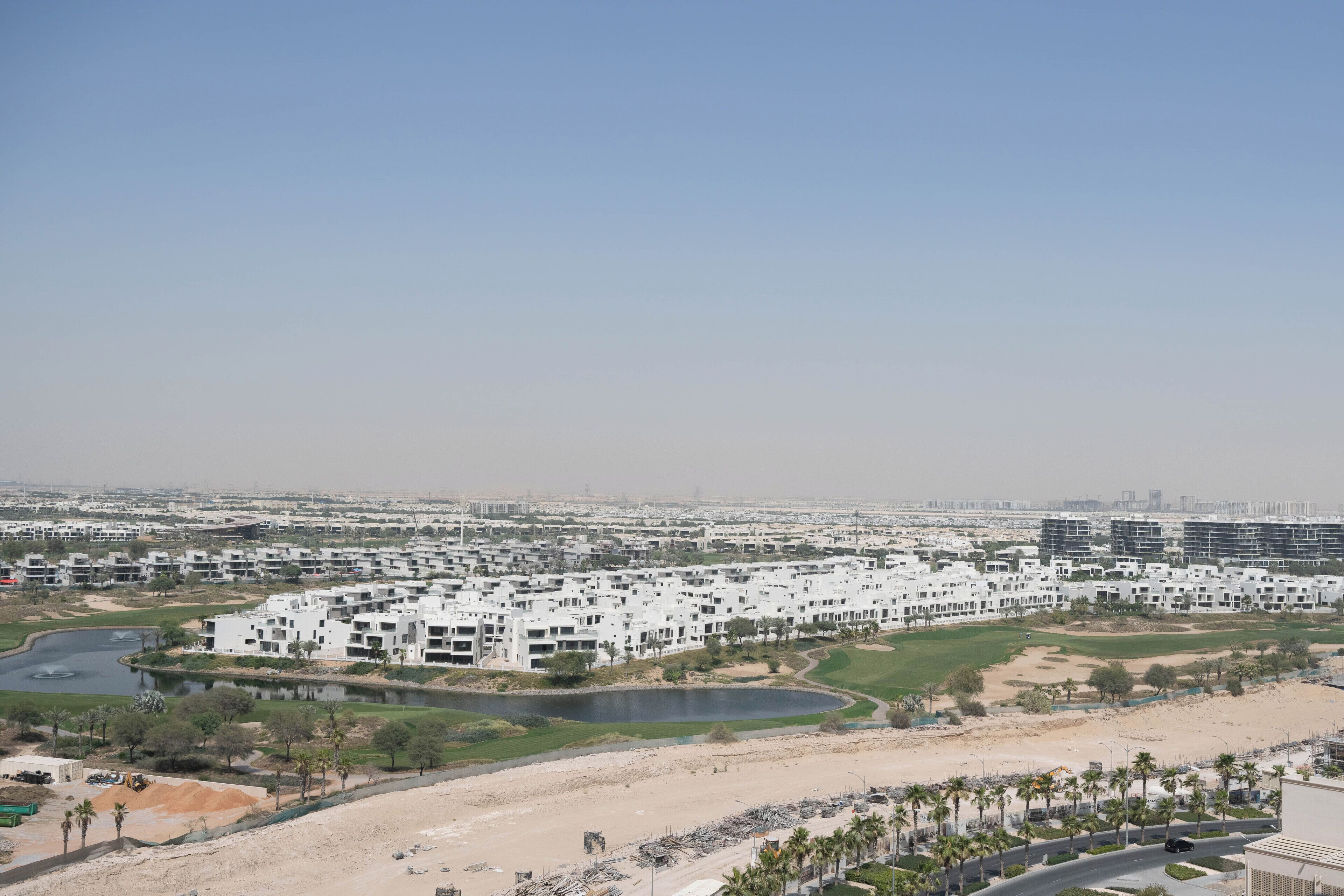 A modern residential complex with multiple buildings, a central water feature, and a sandy area with palm trees.