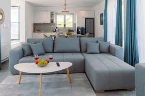Ein modernes Wohnzimmer mit einem grauen Ecksofa, einem weißen Couchtisch und einer Küche im Hintergrund.
