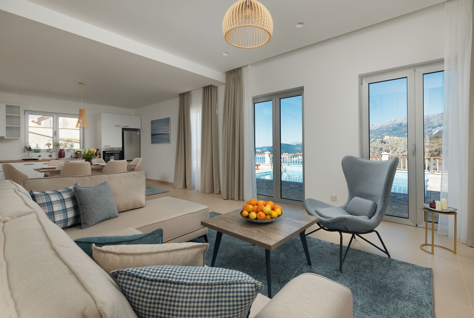 A modern living room with a sofa, armchairs, a coffee table with fruit, and a balcony with a pool view.