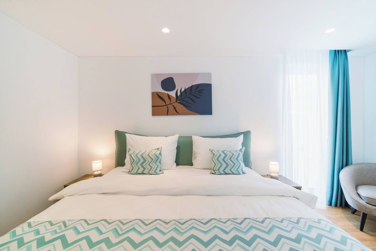 A modern hotel room with a bed, two bedside lamps, a wall-mounted abstract art piece, and a view of a blue curtain.