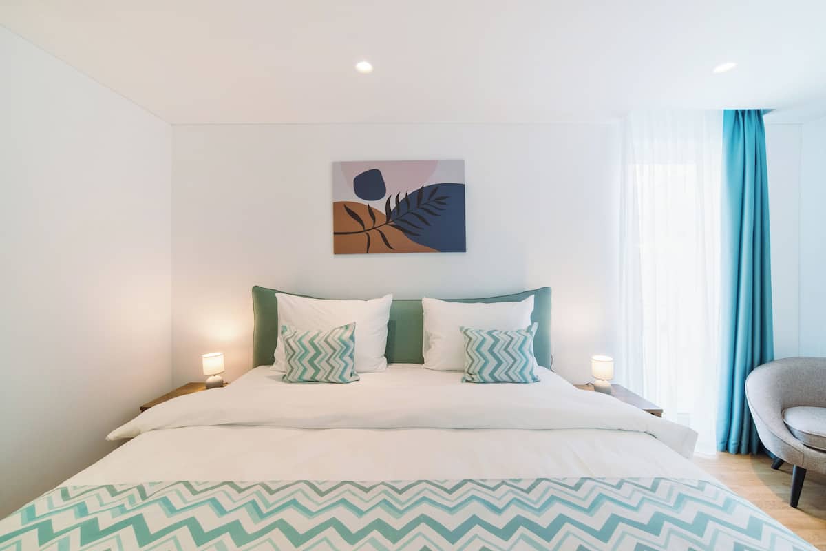 A modern hotel room with a bed, two bedside lamps, a wall-mounted artwork, and a window with curtains.