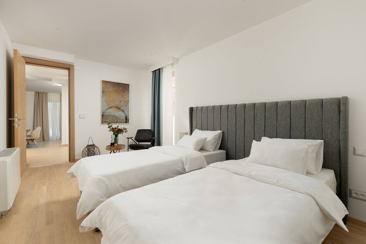 A hotel room with two beds, a sitting area with a chair and a small table, and a view of the hallway.