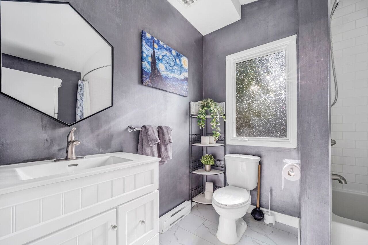 A bathroom with a white sink, a toilet, a shower, a large mirror, a painting on the wall, and a window.