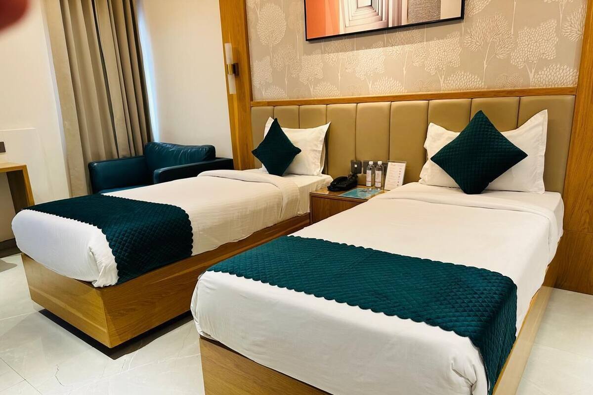 Habitación de hotel con dos camas, cada una con cabecero de madera, ropa de cama blanca y cojines decorativos color turquesa. Se ve una mesita de noche con una lámpara y una pequeña mesa con un libro y un bloc de notas.