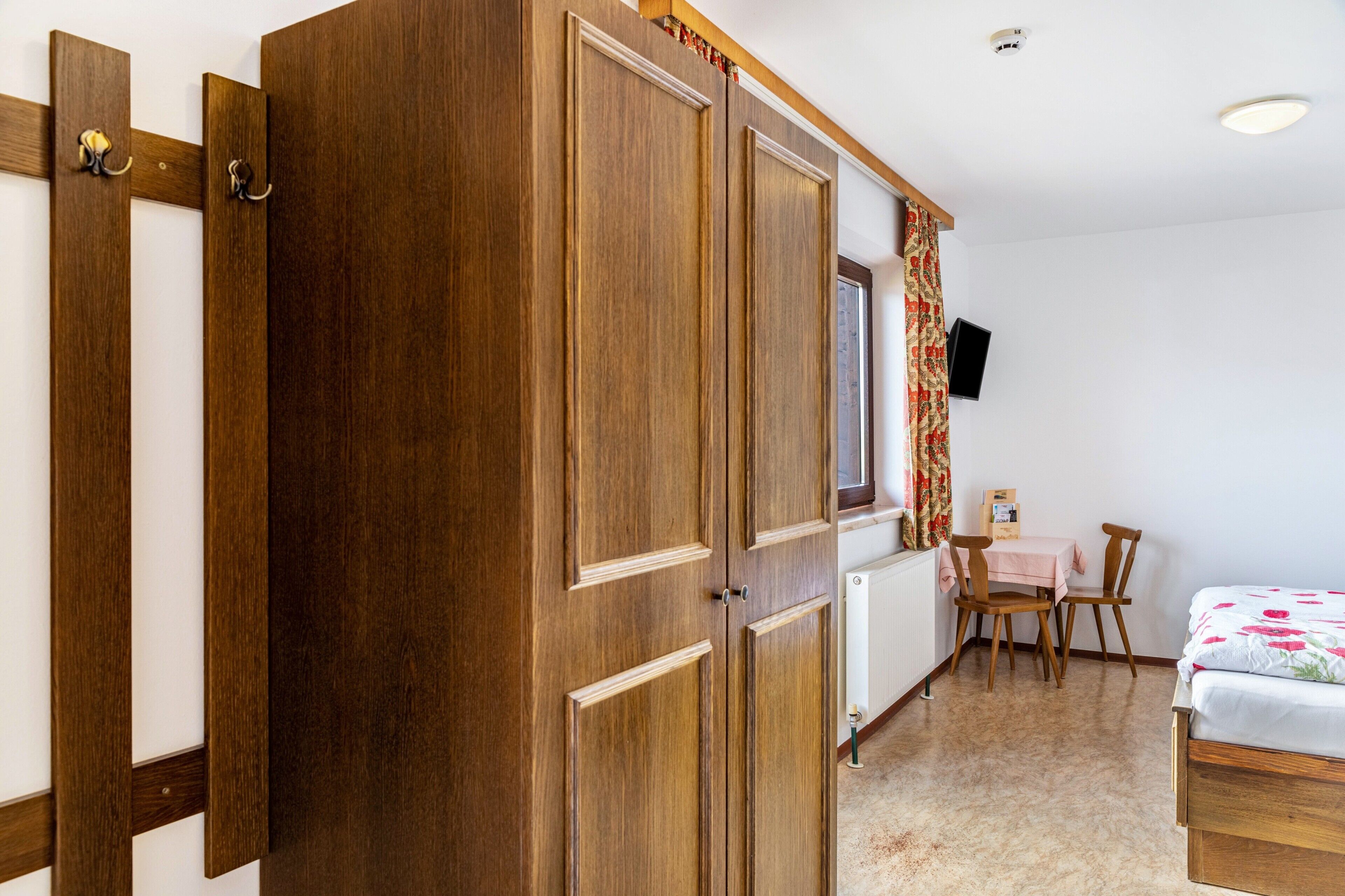 Een slaapkamer met een houten garderobe, een bed, een bureau met stoelen, een radiator en een televisie aan de muur.