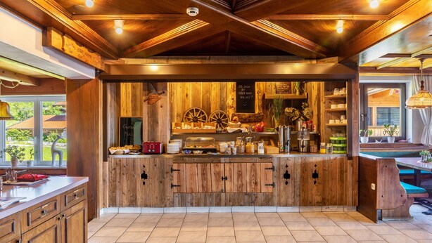 Une cuisine rustique avec des meubles en bois, un îlot central où sont disposés des produits alimentaires et un tableau noir servant de menu.