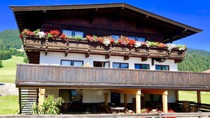 Un chalet en bois à deux étages, doté de balcons ornés de fleurs, d’un espace extérieur couvert aménagé en salon et offrant une vue sur une vallée verdoyante.