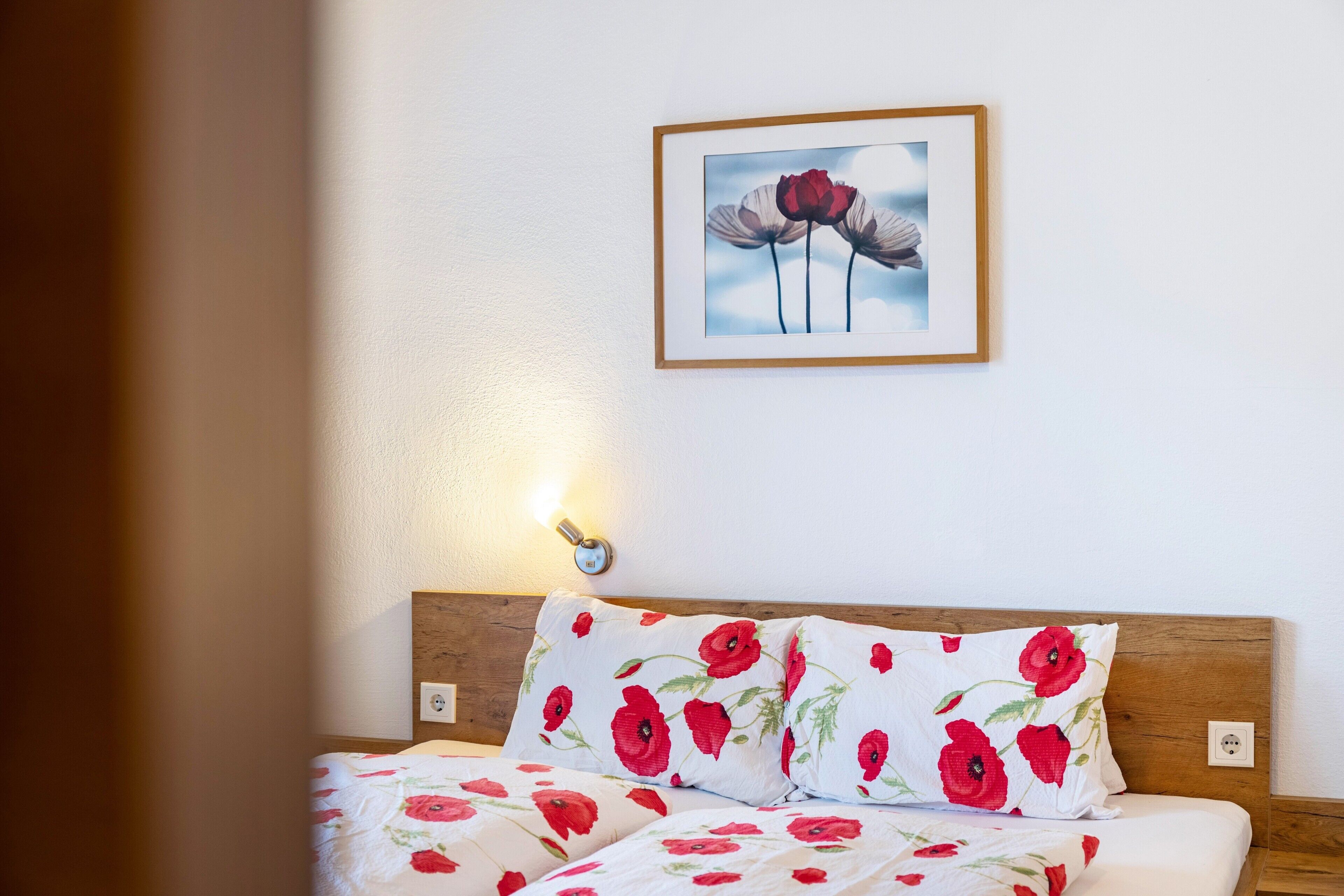 Cama bem arrumada com roupa de cama florida, um quadro na parede e uma luminária fixada na parede.