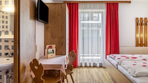 Ein Schlafzimmer mit einem hölzernen Bett, einem Nachttisch, einem Sessel, einem an der Wand befestigten Fernseher und einem Fenster mit roten Vorhängen.