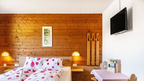 Un dormitorio con pared de madera, una cama con ropa de cama floral, lámparas de noche, un televisor y una pequeña mesa con sillas.