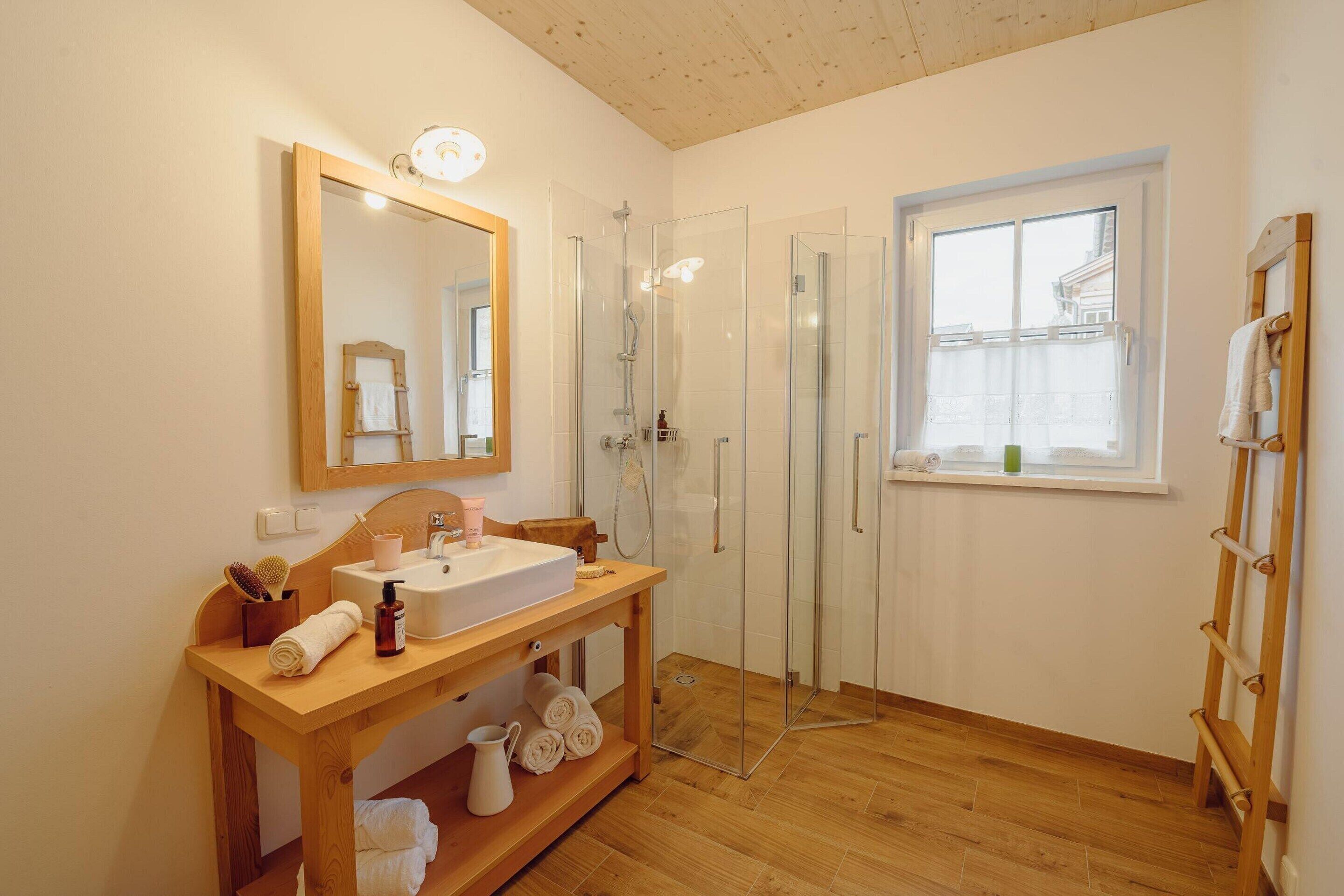 Une salle de bain avec un meuble-lavabo en bois, une cabine de douche en verre, un grand miroir et une fenêtre donnant sur des immeubles.