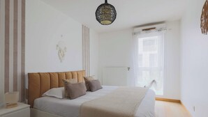 Une chambre à coucher comprenant un lit, des tables de chevet, un luminaire suspendu, une fenêtre avec des rideaux et un radiateur.
