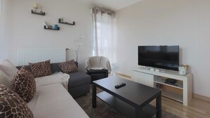 Un salon comprenant un canapé, une table basse, un téléviseur et un fauteuil. Des étagères ornées d’objets décoratifs sont fixées au mur.