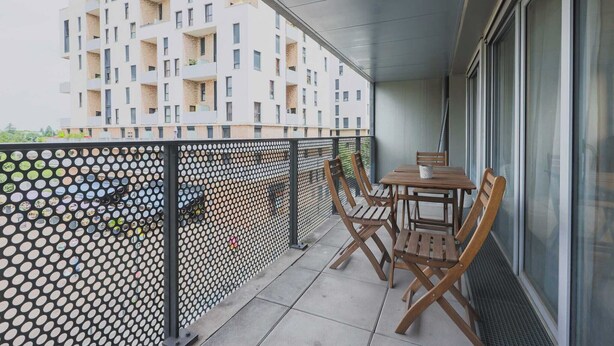 Un balcon avec une table et des chaises, une clôture en métal ajouré et, au fond, un immeuble doté de nombreux balcons.