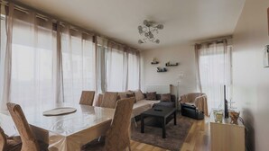 Un salon moderne comprenant un coin repas, de grandes fenêtres avec des rideaux légers, un luminaire central et une petite table d’appoint agrémentée d’objets décoratifs.
