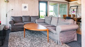 Un salon chaleureux avec un plafond en bois, des canapés gris, une table basse ronde en bois et une grande fenêtre donnant sur l’extérieur.