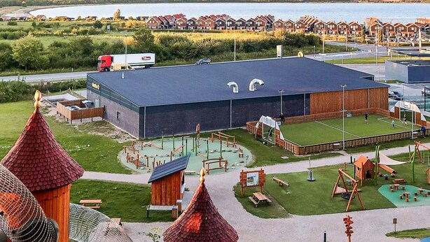 Un grand bâtiment doté d’un toit plat, entouré d’une aire de jeux avec différents équipements, et avec une étendue d’eau en arrière-plan.