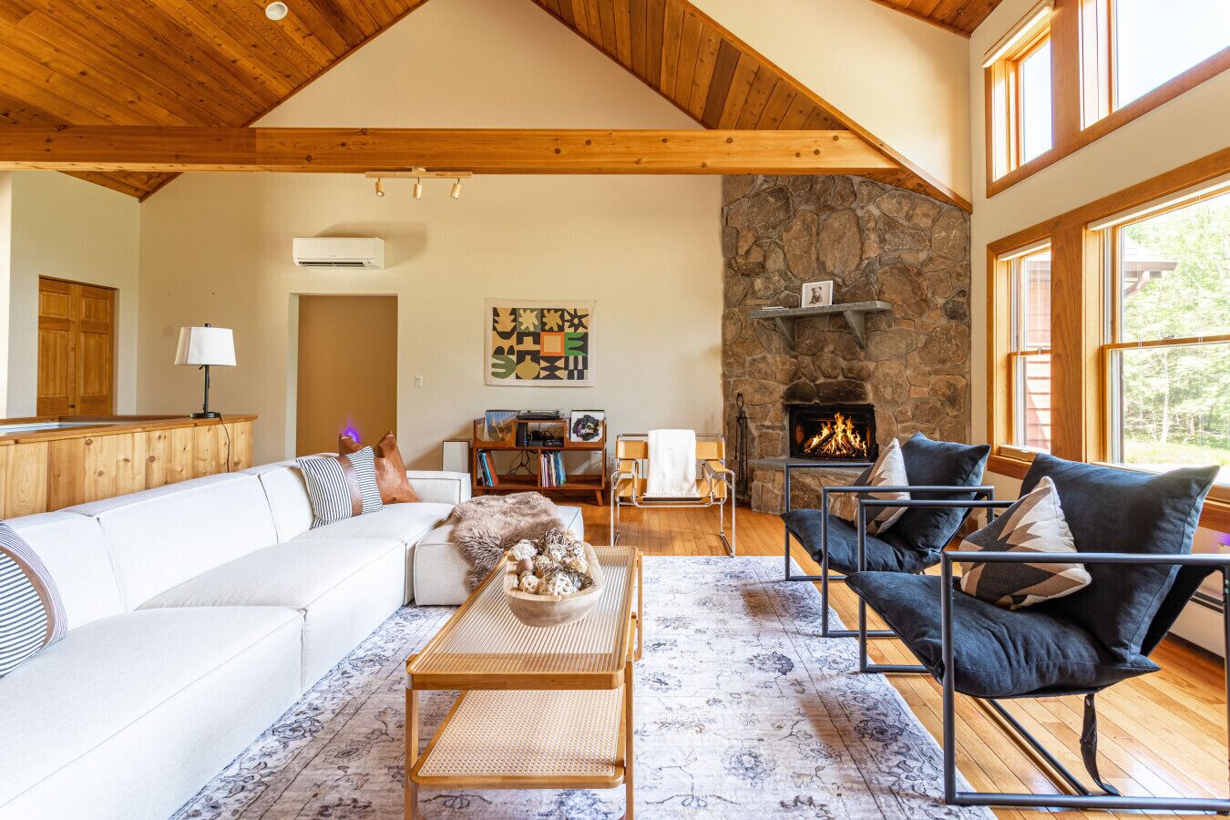 A spacious living room with a stone fireplace, wooden ceiling beams, and large windows.
