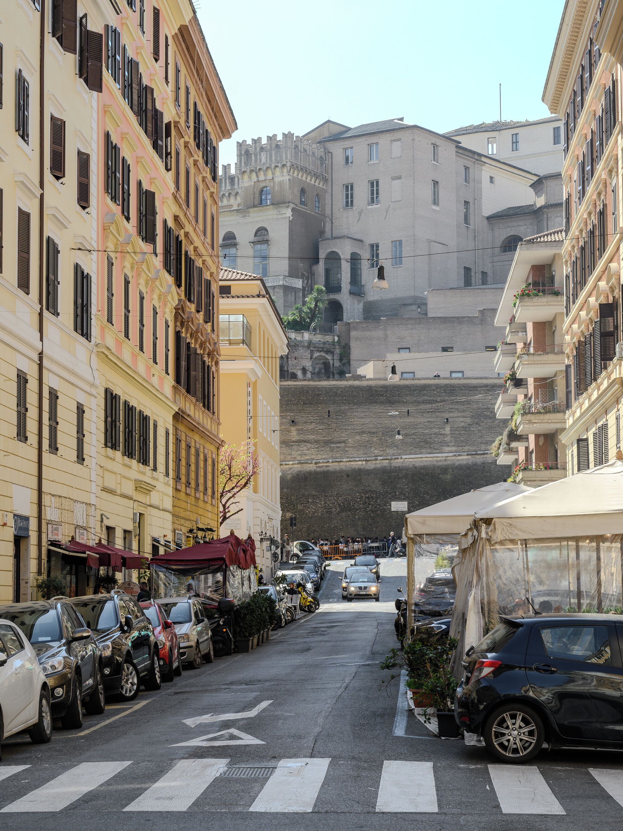 A narrow street lined with multi-story buildings, parked cars, and a few outdoor seating areas.