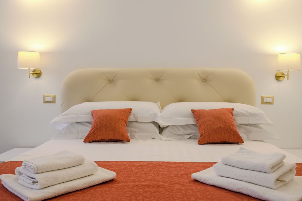 A neatly made bed with white linens and two orange pillows, a red accent rug, and two wall-mounted lamps.