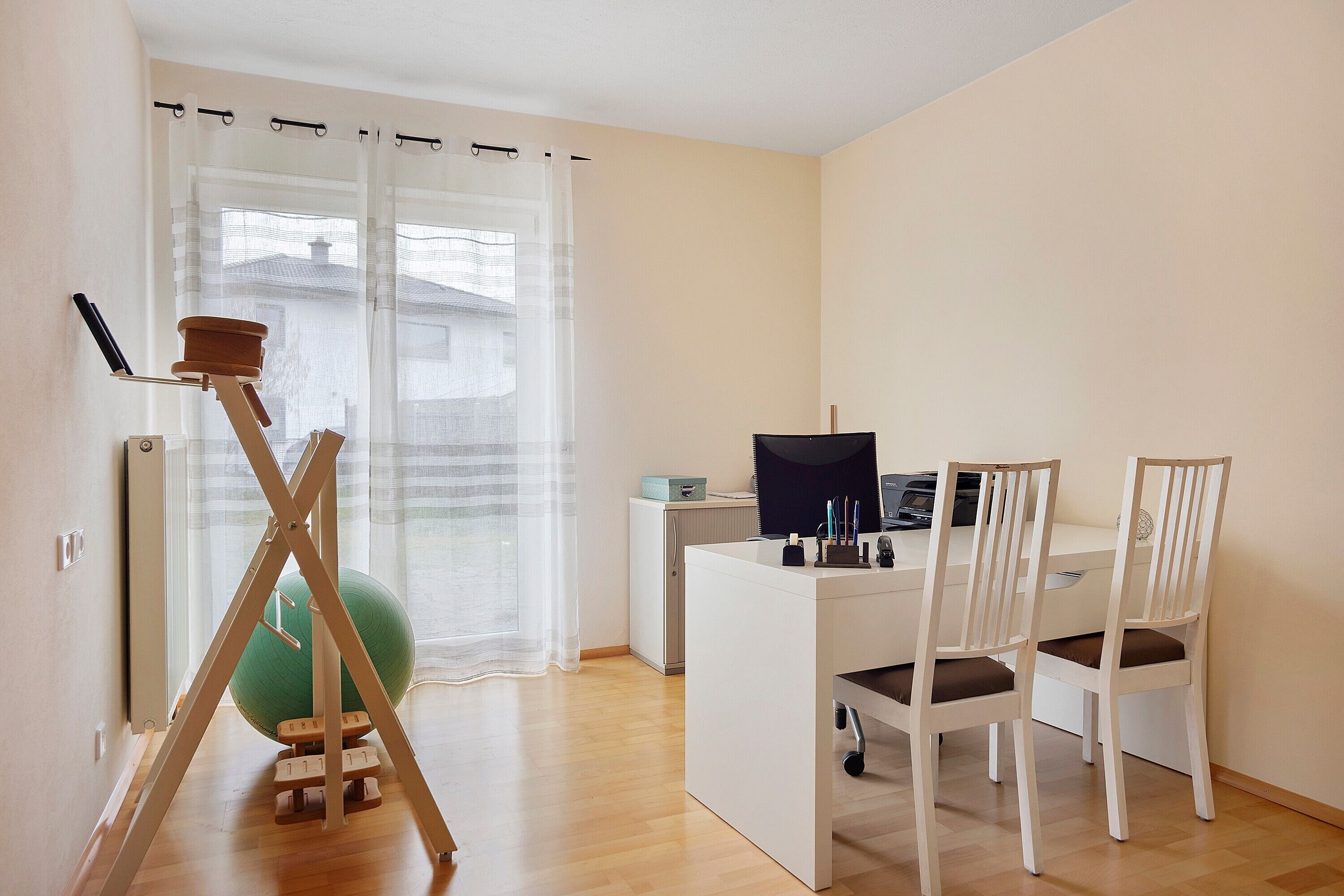 Ein Zimmer mit Schreibtisch, zwei Stühlen, einem Computer, einem grünen Gymnastikball und einem Stehtisch mit Sitzmöglichkeit.