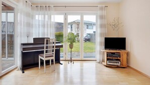 Una habitación con un piano, un televisor sobre un soporte de madera, una planta y un amplio ventanal corredero.
