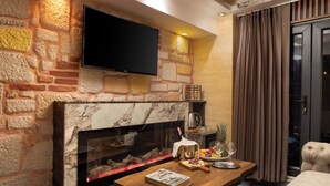 A living room with a stone wall, a flat-screen TV mounted above a fireplace, a coffee table with a book, a fruit bowl, and a pineapple. - REN PLUS SUİTES OTEL (İstanbul)
