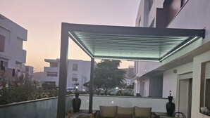En takterrass med en pergola av glas, en sittgrupp och krukväxter.