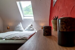 Une chambre avec un plafond en pente, un lit recouvert d’une literie blanche, une petite table avec une tasse, et une fenêtre donnant sur un paysage verdoyant.