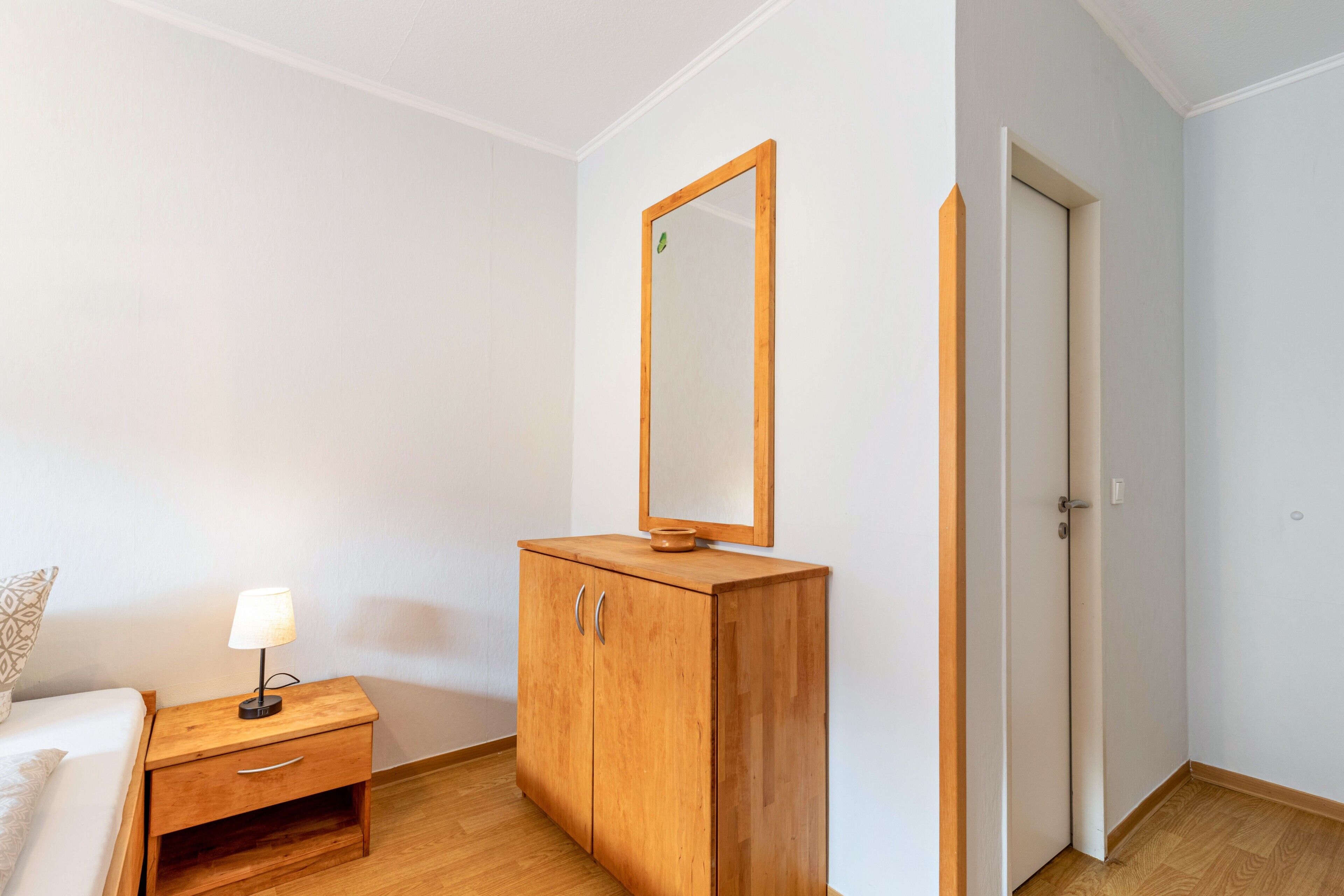 Ein Schlafzimmer mit einem Holzschrank, einem Nachttisch mit Lampe, einem Spiegel und einer Tür.