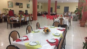 Un comedor con varias mesas dispuestas para comidas, con manteles blancos, servilletas rojas y un frutero central.