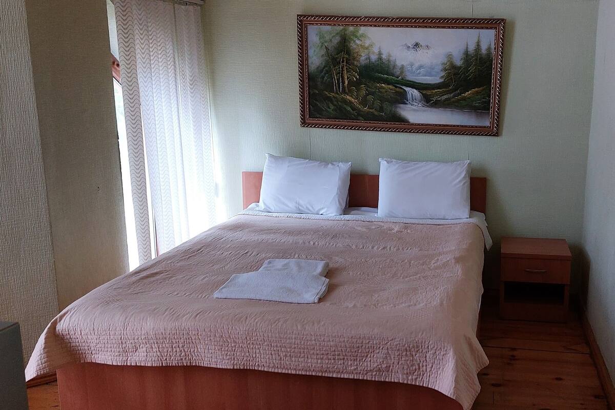 Un lit bien fait, avec une couvre-lit rose et des oreillers blancs, une petite table de chevet et une commode. Un tableau représentant une scène de forêt est accroché au-dessus du lit.