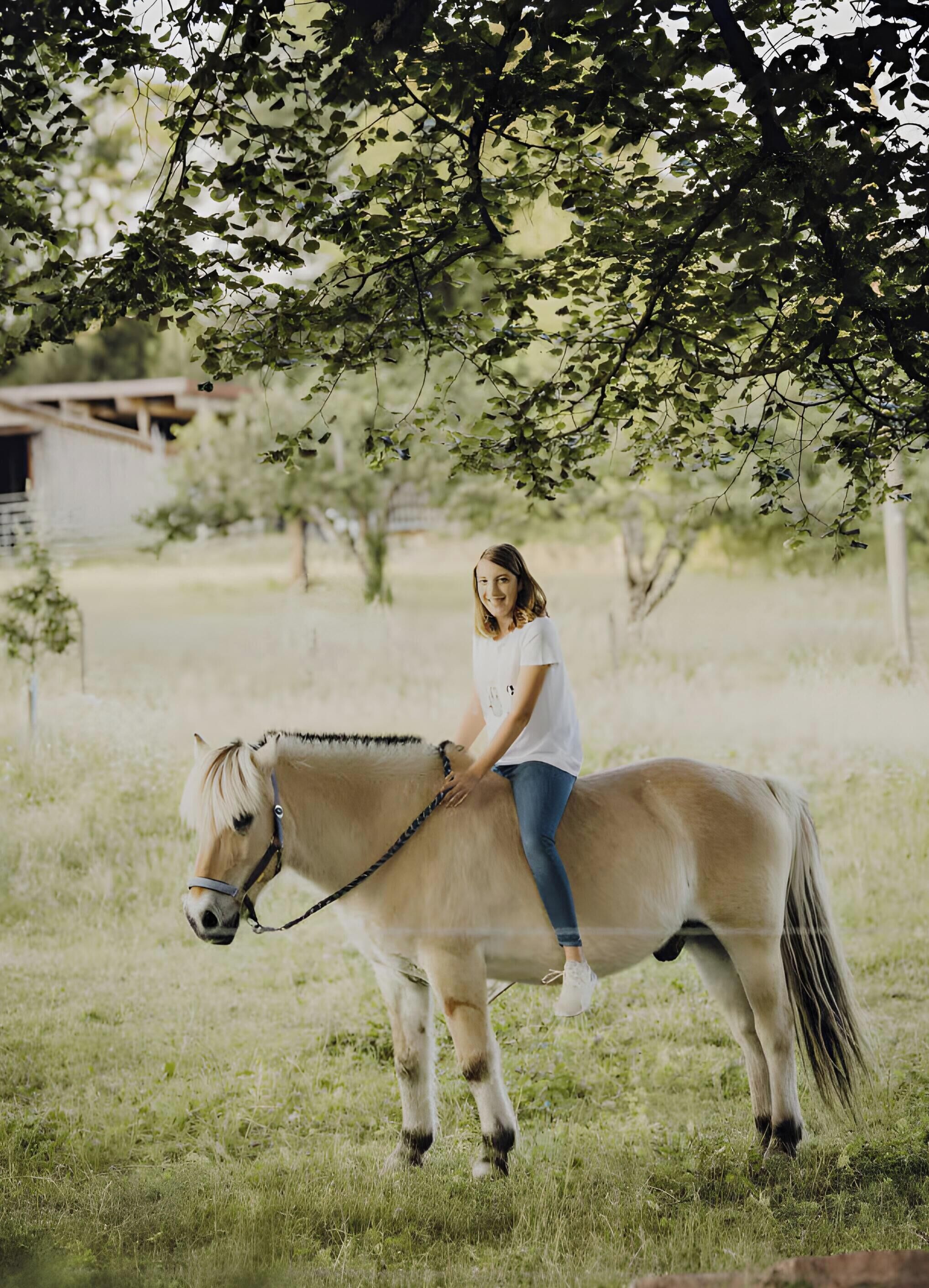 A person riding a horse in a grassy field with trees and a building in the background.