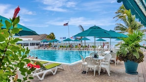 Área de piscina com espreguiçadeiras e guarda-sóis, uma bandeira e vista para o mar.