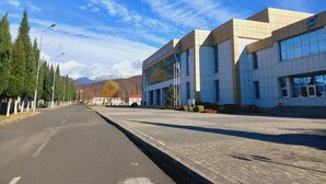 Un edificio moderno con facciata in vetro, una strada pavimentata alberata e montagne sullo sfondo.