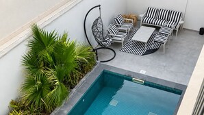 Una piscina rectangular con borde de azulejos, rodeada por un jardín con follaje verde y un área de descanso con cojines a rayas y una silla colgante.