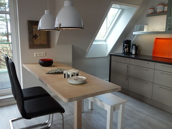 A modern kitchen with a wooden table, two pendant lights, a black chair, and a window with a view.