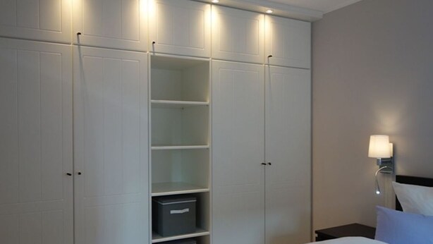 A bedroom with a bed, a wardrobe with shelves, and a nightstand with a lamp.