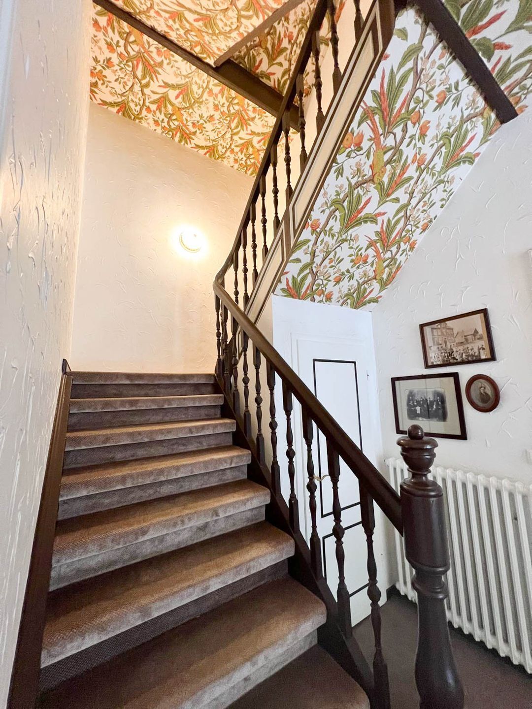 A staircase with wooden steps and a dark wooden handrail, leading to a landing with a floral-patterned wallpaper and a white door.