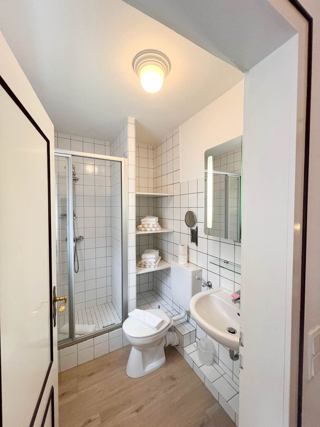 Ein Badezimmer mit Dusche, Toilette und Waschbecken, mit weißen Fliesen und Holzboden.