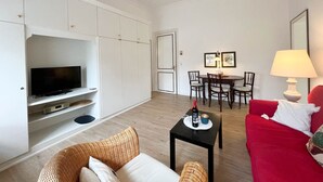 A living room with a TV, a red sofa, a wicker chair, a small table with a bottle and glasses, and a dining area with chairs and a table. - House Friesland - Apartment 2 - An urban villa with spacious and tasteful (Sylt)