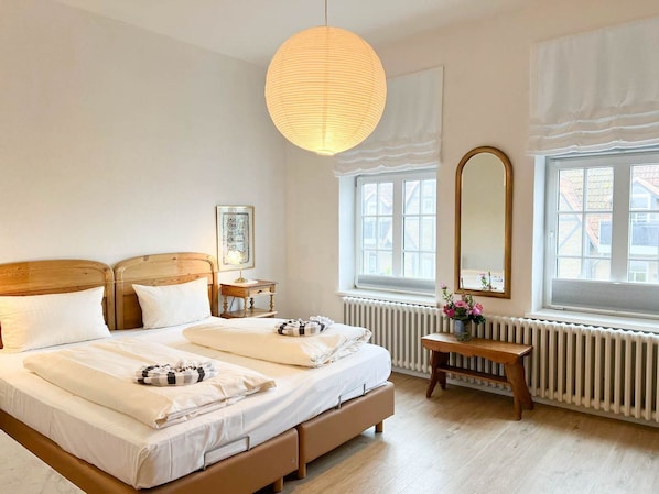 Un lit double avec du linge de lit blanc, une tête de lit en bois, une table de chevet avec une lampe, une grande fenêtre donnant sur un beau paysage et un radiateur.