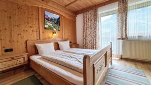 Une chambre à coucher dans un chalet en bois, avec un lit, une table de chevet, une lampe fixée au mur, une fenêtre avec des rideaux et une vue sur l’extérieur.