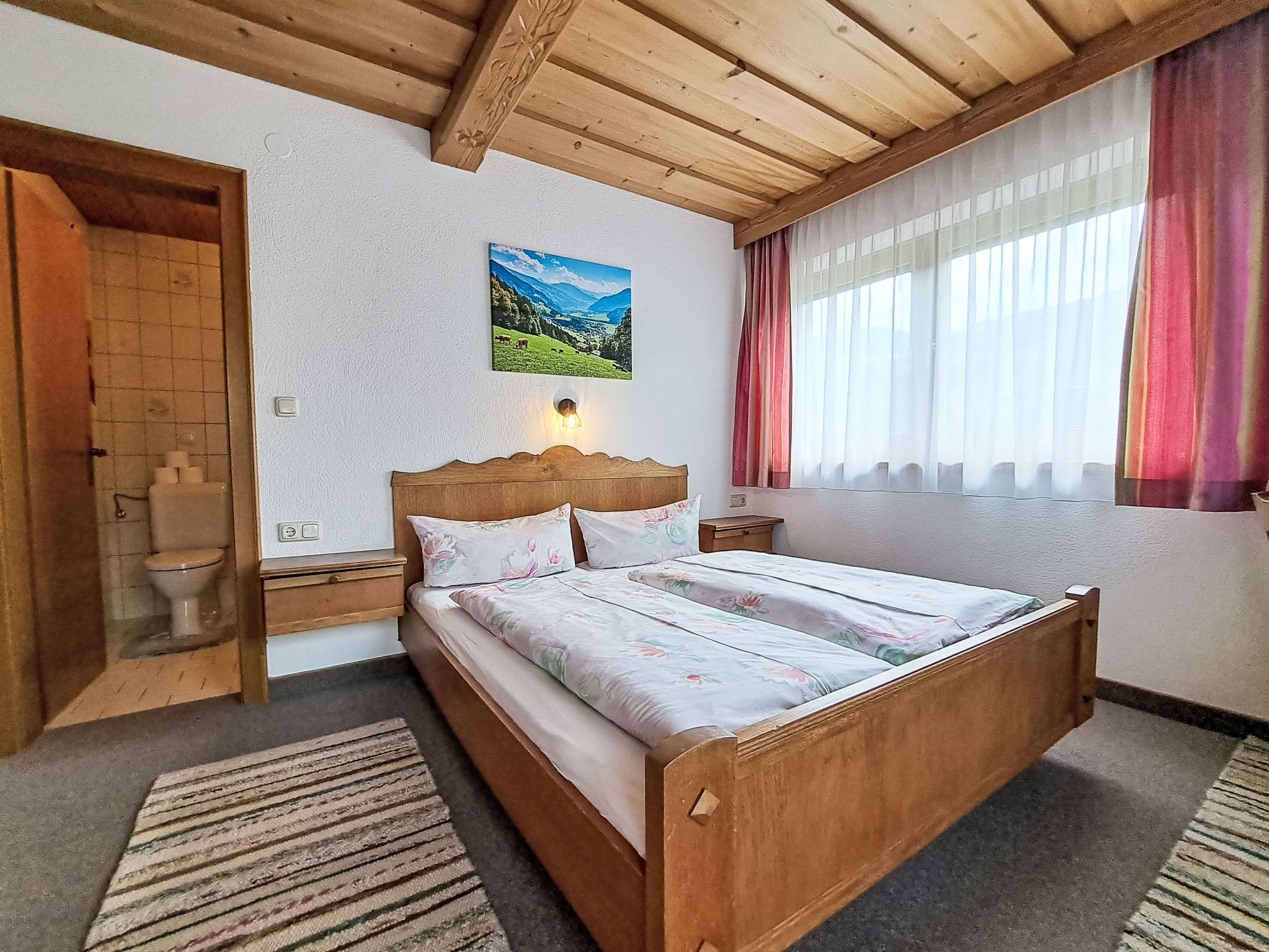 Een slaapkamer met een houten bed, een nachtkastje, een wandlamp, een schilderij van een landschap, en een badkamer met toilet en douche.